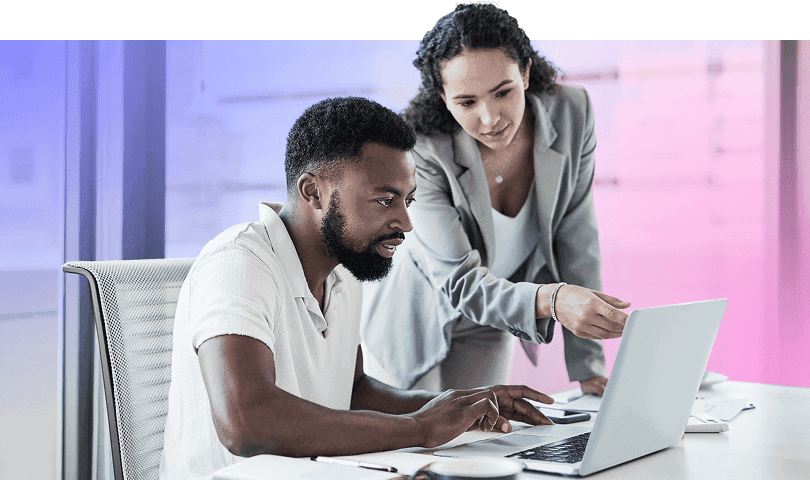 Two professionals collaborating at a desk, with one person typing on a laptop and the other pointing at the screen.
